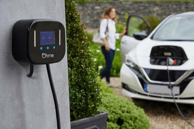 Ohme Home Pro laadpaal aan de muur, op de achtergrond een vrouw die naar haar auto loopt